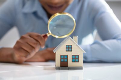 Close-up Of A Businesswoman's Hand Holding Magnifying Glass Over House Model Over Desk